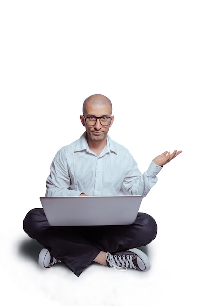 Retrato de um jovem latino na camisa branca e calça preta segurando o laptop enquanto está sentado no chão e apontando com a mão isolada sobre fundo branco