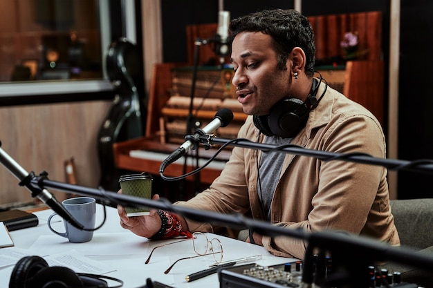 Retrato de um jovem locutor de rádio tomando uma bebida, parecendo focado enquanto fala no microfone