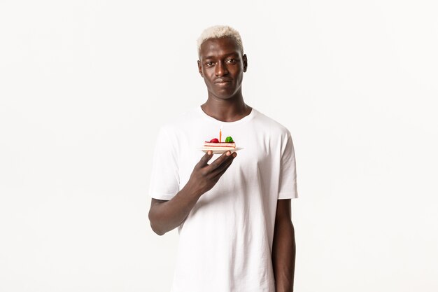 Retrato de um jovem loiro afro-americano sombrio, segurando um bolo de aniversário com uma vela e parecendo relutante