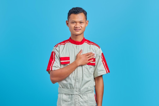 Retrato de um jovem mecânico asiático sorridente com as palmas das mãos no peito, sobre um fundo azul
