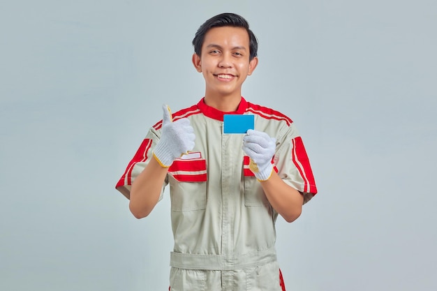 Retrato de um jovem mecânico bonito mostrando o cartão de crédito na mão e o polegar para cima gesto no fundo cinza