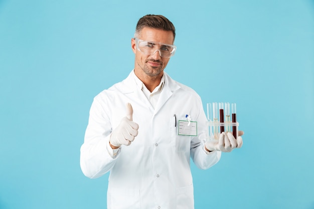 Retrato de um jovem médico de óculos segurando tubos de ensaio com sangue, isolado sobre a parede azul