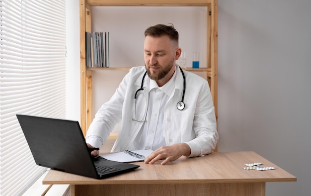 Retrato de um jovem médico em um casaco branco com um estetoscópio sentado em um consultório médico e trabalhando em um laptop