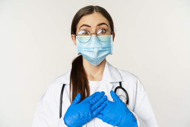 Retrato de um jovem médico surpreso e espantado, olhando com fascínio, de mãos dadas no coração, de pé com máscara facial médica sobre fundo branco.