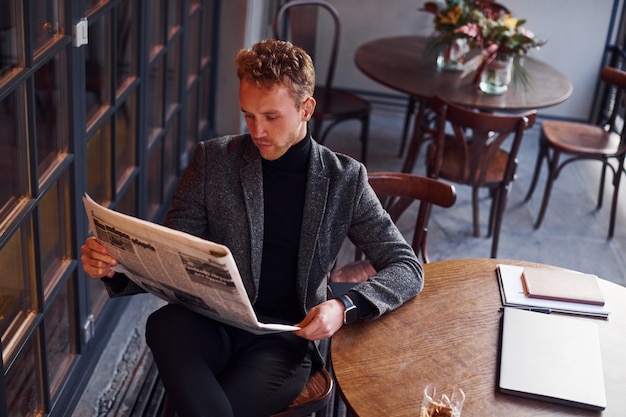 Retrato de um jovem moderno com roupas formais, que se senta no café e lê o jornal.