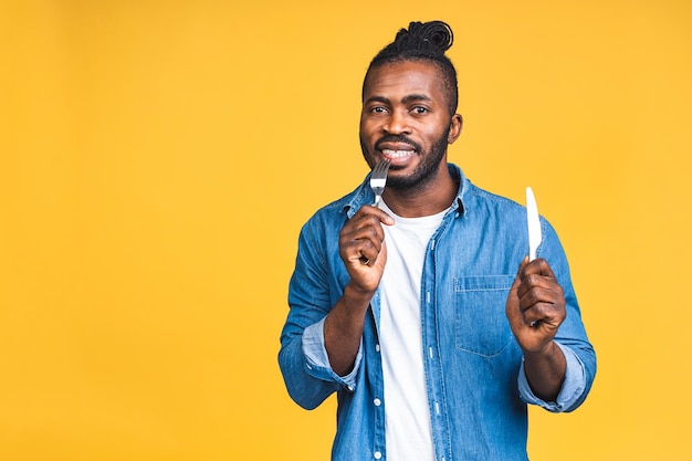 Retrato de um jovem negro afro-americano alegre feliz sorridente segurando colher e faca em pé isolado sobre fundo amarelo.