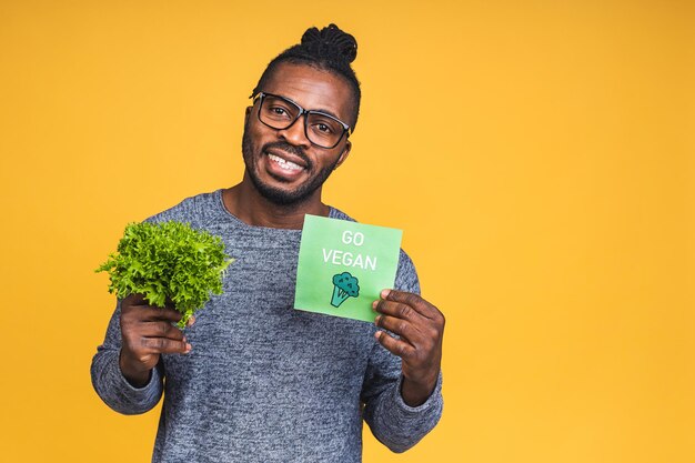 Retrato de um jovem negro afro-americano bonito segurando folhas de alface frescas isoladas sobre fundo amarelo. Conceito de estilo de vida vegan.