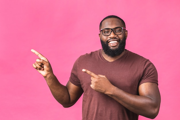 Retrato de um jovem negro afro-americano feliz com um sorriso casual, apontando para o lado com o dedo, olhando na câmera com expressão de rosto animado isolada sobre fundo rosa.