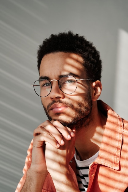 Retrato de um jovem negro carrancudo em óculos redondos posando na sombra das cortinas