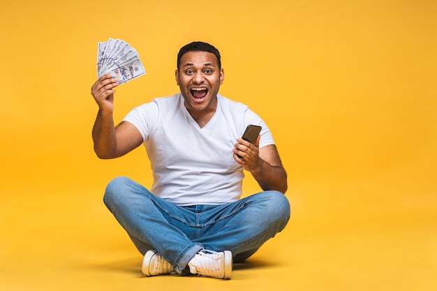 Retrato de um jovem negro indiano afro-americano, sentado no chão, segurando as notas de dólar isoladas sobre fundo amarelo. Usando o celular.