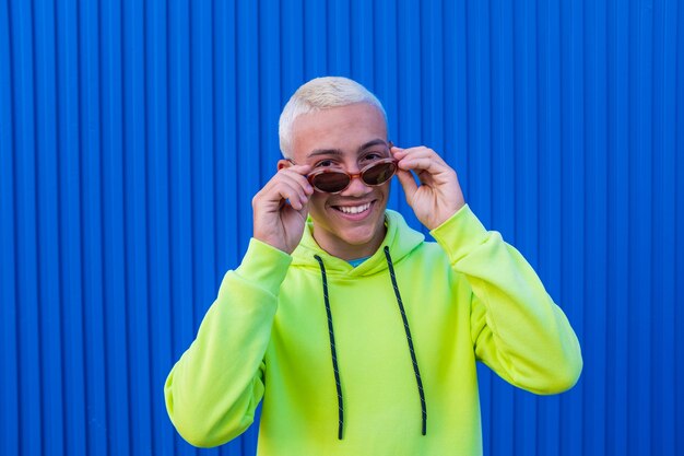 Retrato de um jovem ou adolescente sorrindo e se divertindo usando óculos de sol da moda e um suéter verde-limão com fundo azul colorido - pessoas alegres do milênio curtindo