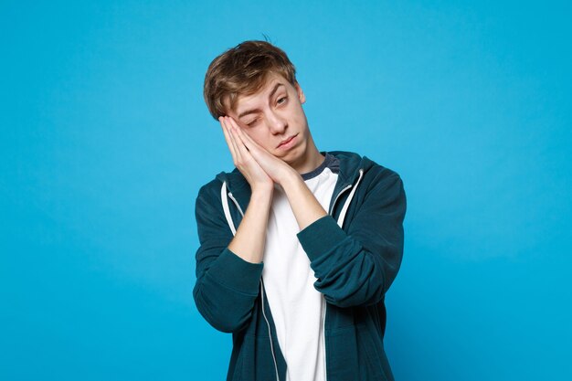 Retrato de um jovem relaxado em roupas casuais, descansando, mantendo um olho fechado, dormindo com as mãos perto do rosto isolado na parede azul. conceito de estilo de vida de emoções sinceras de pessoas.