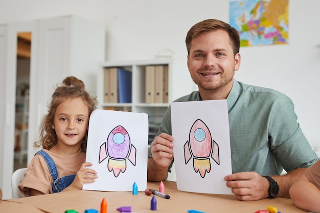 Retrato de um jovem sorridente e uma menina mostrando fotos de foguetes espaciais enquanto desfrutavam da aula de arte na pré-escola juntos