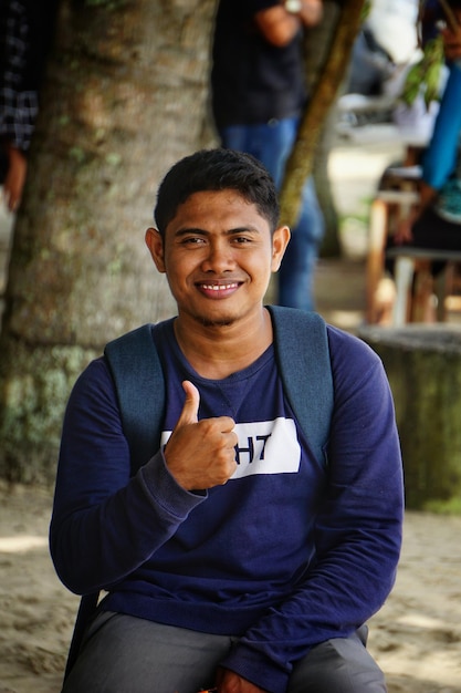 Foto retrato de um jovem sorridente mostrando os polegares contra a árvore