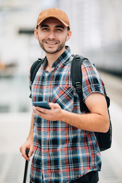 Retrato de um jovem sorridente sentado com um telefone celular e uma bolsa esperando o trem na estação