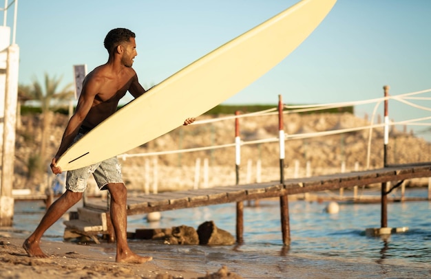 Retrato de um jovem surfista negro profissional segura sua prancha de surf no braço fica no sol do sol da praia do oceano atrás do ombro esporte hobby estilo de vida relaxado