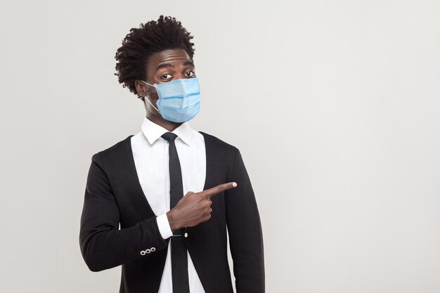 Retrato de um jovem trabalhador bonito e engraçado vestindo terno preto com máscara médica cirúrgica em pé mostrando e apontando para o espaço vazio de fundo. tiro de estúdio interior isolado em fundo cinza.