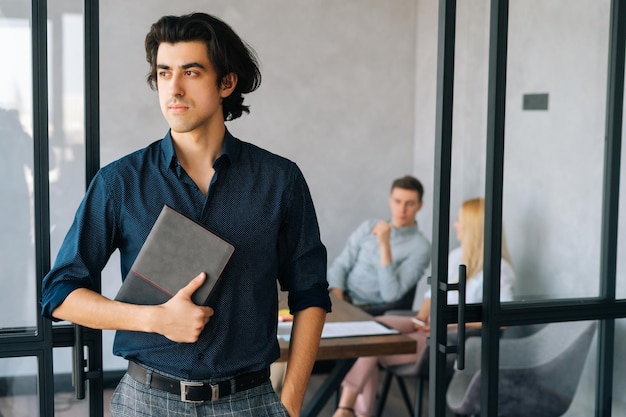 Retrato de um líder de equipe sério segurando na mão um elegante caderno de papel parado em uma sala de escritório moderna pensativo olhando para longe