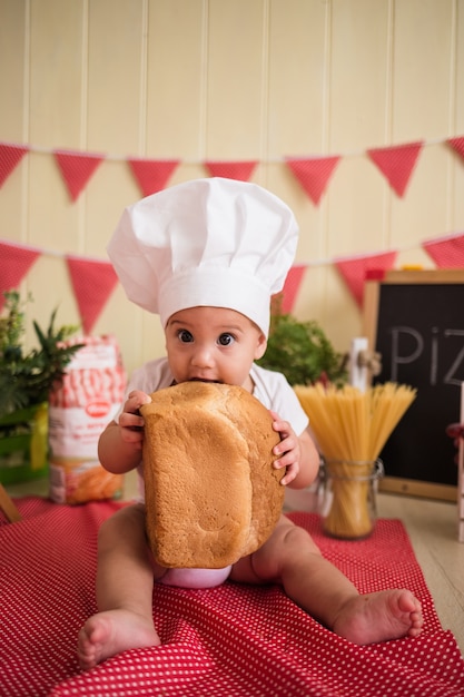 Retrato de um lindo bebê com um chapéu branco de chef comendo pão