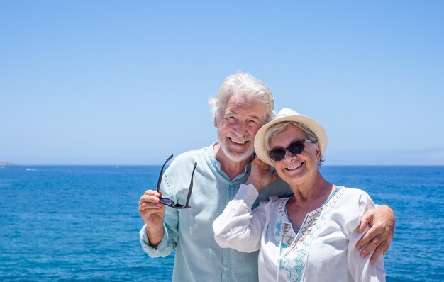 Retrato de um lindo casal sênior abraçado em frente ao mar, olhando para a câmera. aposentados felizes em pé na praia aproveitando as férias de verão