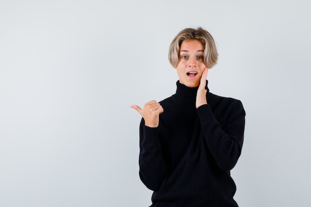 Retrato de um lindo menino adolescente apontando para a esquerda com o polegar, com a mão na bochecha, em um suéter preto de gola alta e olhando maravilhado com a vista frontal