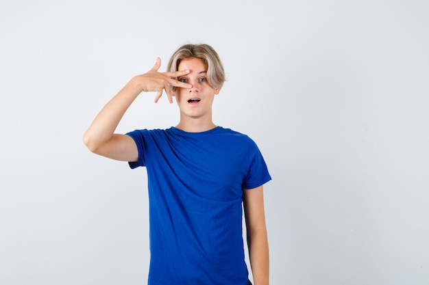 Retrato de um lindo menino adolescente mostrando o sinal de V no olho em uma camiseta azul e olhando maravilhado para a vista frontal