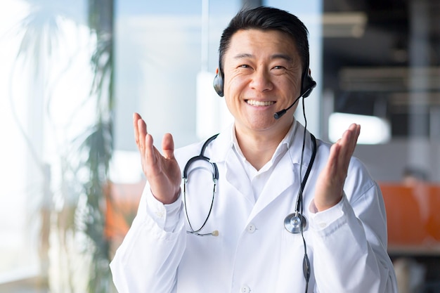 Retrato de um médico asiático experiente olhando para a câmera e se divertindo conversando com o paciente sorrindo amigavelmente e regozijando-se com a recuperação do paciente trabalhando remotamente videochamada com fone de ouvido