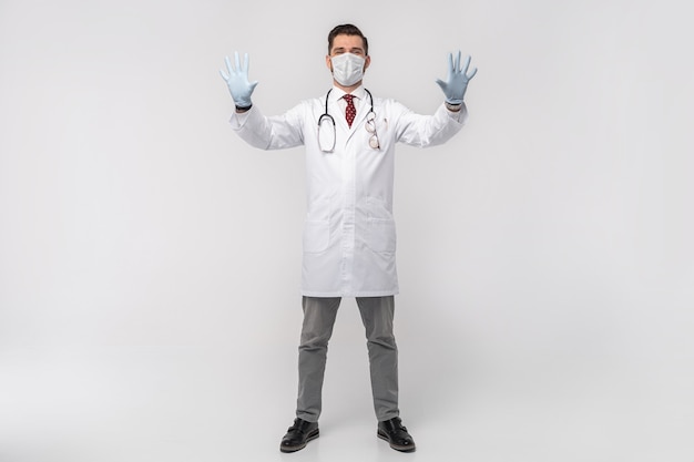 Retrato de um médico bonito atraente em máscara protetora, jaleco branco, gravata isolada na parede branca