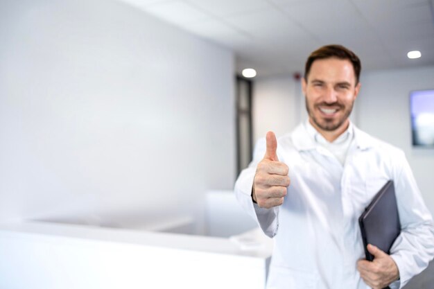 Foto retrato de um médico caucasiano sorridente segurando um computador portátil dentro do lobby do hospital