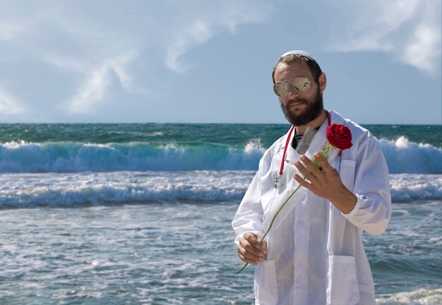 Retrato de um médico judeu barbudo em yarmulke branco (chapéu, kipá, chapéu judeu) usando óculos escuros, casaco e estetoscópio segurando uma flor rosa vermelha na mão. homem barbudo bonito americano no fundo do mar