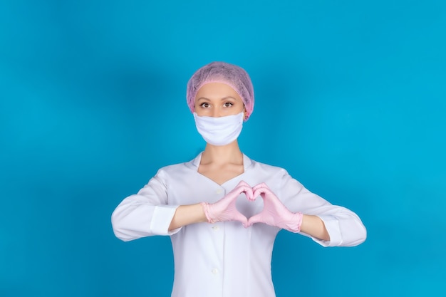 Retrato de um médico ou enfermeiro em uma máscara médica e luvas de nitrilo rosa, mãos mostrando um coração saudável, isolado em uma parede azul.