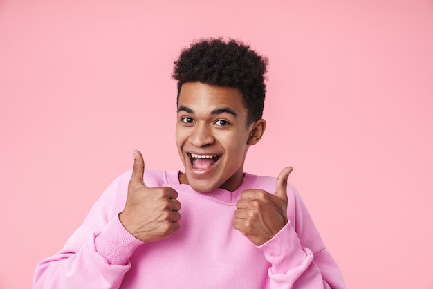 Retrato de um menino adolescente africano sorridente usando pullower em pé isolado na rosa, dando sinal de positivo