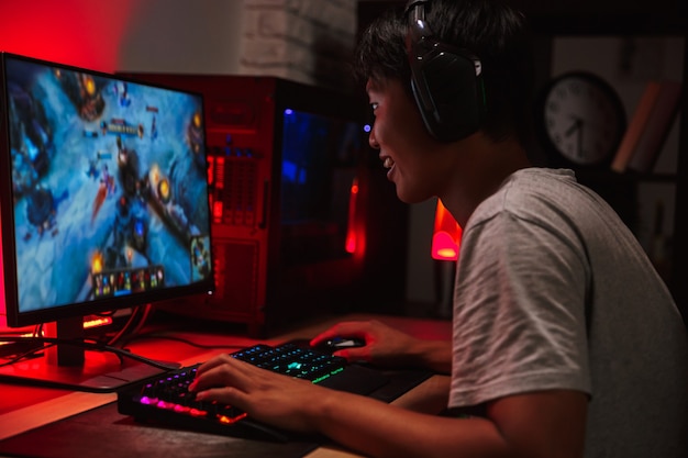 Retrato de um menino adolescente asiático feliz jogando videogame no computador no quarto escuro, usando fones de ouvido e um teclado colorido com luz de fundo