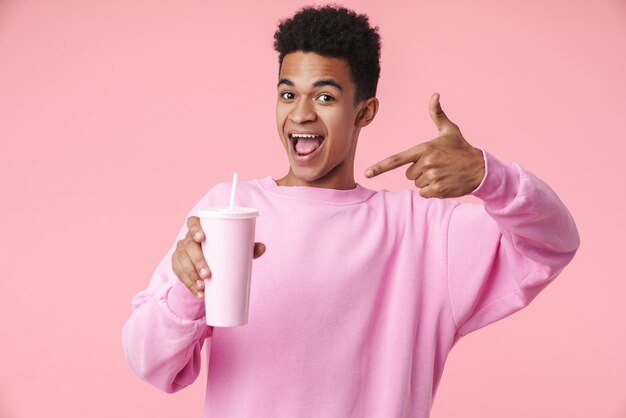 Retrato de um menino adolescente sorridente, usando um puxador isolado na parede rosa, apresentando o copo de plástico com um canudo