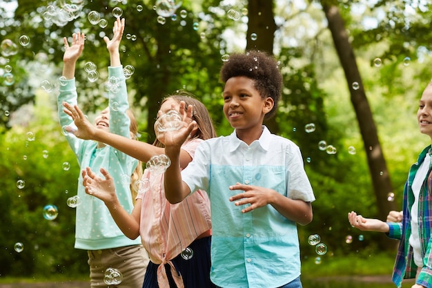 Retrato de um menino afro-americano e um grupo de crianças despreocupadas correndo no parque enquanto brinca com bolhas ao ar livre