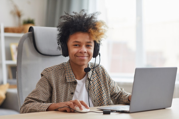 Retrato de um menino afro-americano sorridente usando fone de ouvido enquanto usa o laptop em casa e olhando para a câmera, copie o espaço