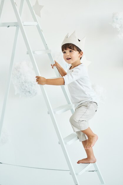 Retrato de um menino bonitinho na coroa na escada branca entre nuvens em um branco