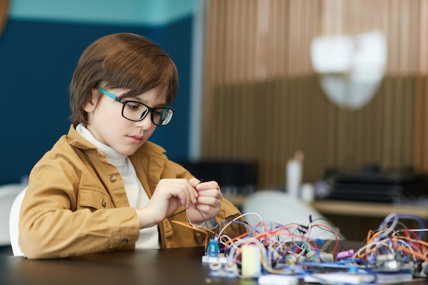 Retrato de um menino bonito usando óculos e trabalhando com fios elétricos enquanto constrói um robô na aula de engenharia, copie o espaço