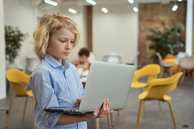 Retrato de um menino calmo olhando focado enquanto segura e usa um laptop em uma sala de aula