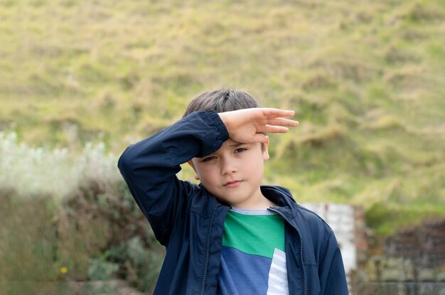 Retrato de um menino com a mão levantada protegendo a luz do sol brilhante brilhando em seus olhos Criança de pé em campos verdes Criança alegre se divertindo brincando ao ar livre na primavera ou no verão