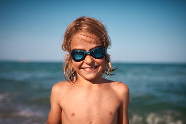Retrato de um menino de 8 anos de cabelos compridos em óculos de natação contra o pano de fundo do mar em uma noite de verão