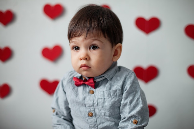 Retrato de um menino de criança em uma camisa com uma gravata borboleta em um fundo de parede com corações no dia dos namorados.