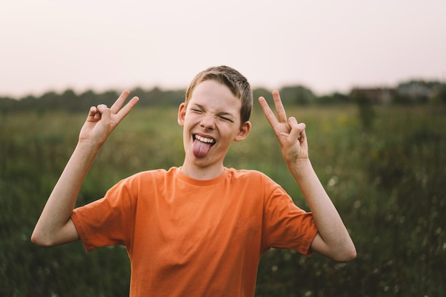 Retrato de um menino engraçado mostra dois dedos vitória ou paz Menino engraçado em uma camiseta laranja brincando ao ar livre no campo ao pôr do sol Estilo de vida de criança feliz