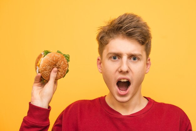 Retrato de um menino expressivo com fome com um hambúrguer na mão em um restaurante amarelo