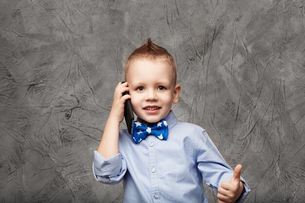 Retrato de um menino fofo com camisa azul e gravata borboleta com telefone celular contra textura cinza em estúdio