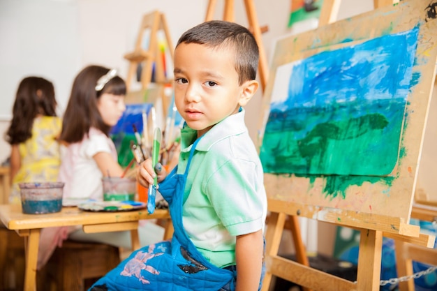 Foto retrato de um menino latino bonitinho vestindo um avental e segurando um pincel, pintando uma paisagem para aula de arte