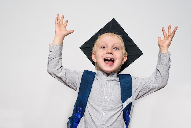 Retrato de um menino loiro alegre com chapéu acadêmico e uma mochila. Mãos ao ar. Fundo branco.