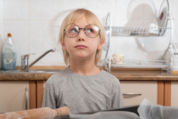 Retrato de um menino loiro com óculos na cozinha. menino pensativo.