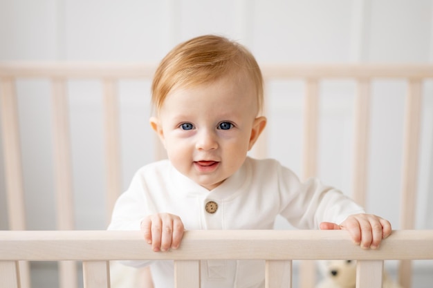 Retrato de um menino loiro de 6 meses de pé em um berço em um quarto infantil brilhante e sorrindo em uma roupa de algodão o conceito de produtos infantis