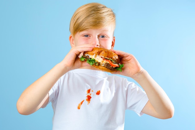 Retrato de um menino mordendo hambúrguer e olhando para a câmera. isolado em fundo azul.
